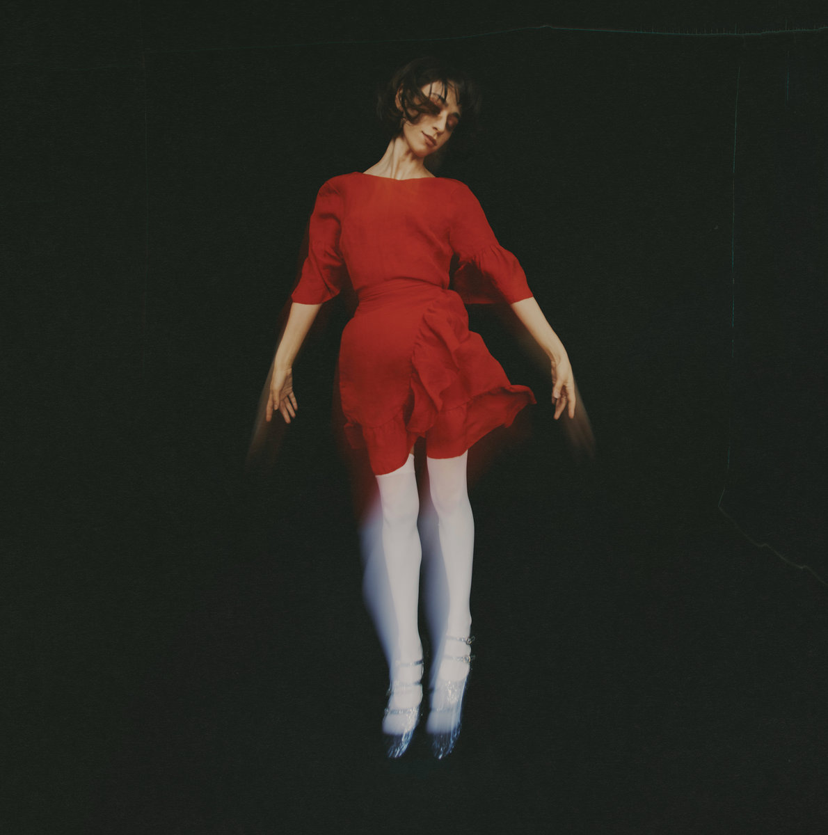 a woman in a red dress, long white socks and silver shoes, the artist Jackie Cohen, appearing to levitate against a black background, or perhaps falling.