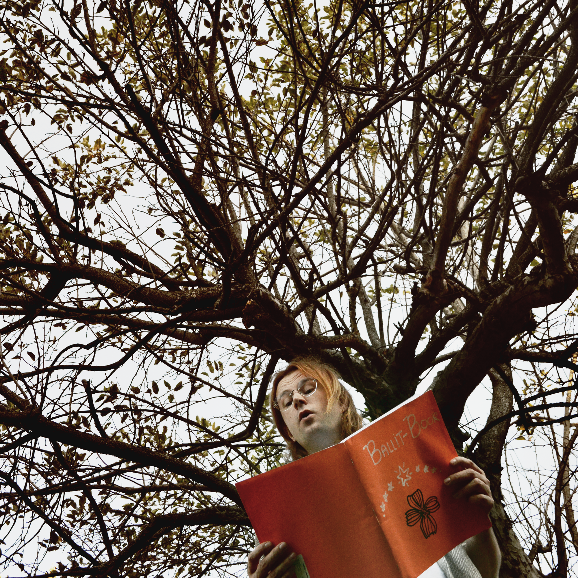 Photo of Derek Piotr reading from a large red book beneath the branches of a tree
