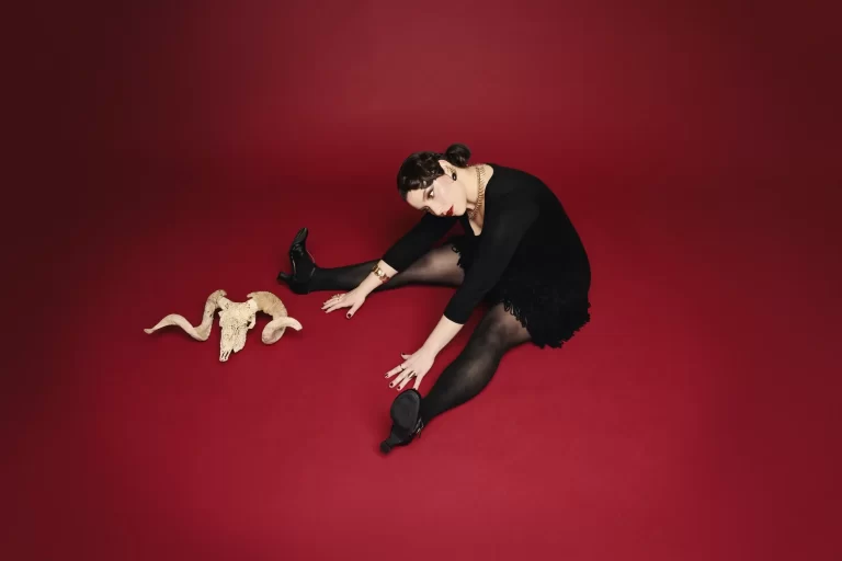 A photo of the artist Gillian Stone showing a woman in a black dress sitting on a red floor with a ram skull beside her