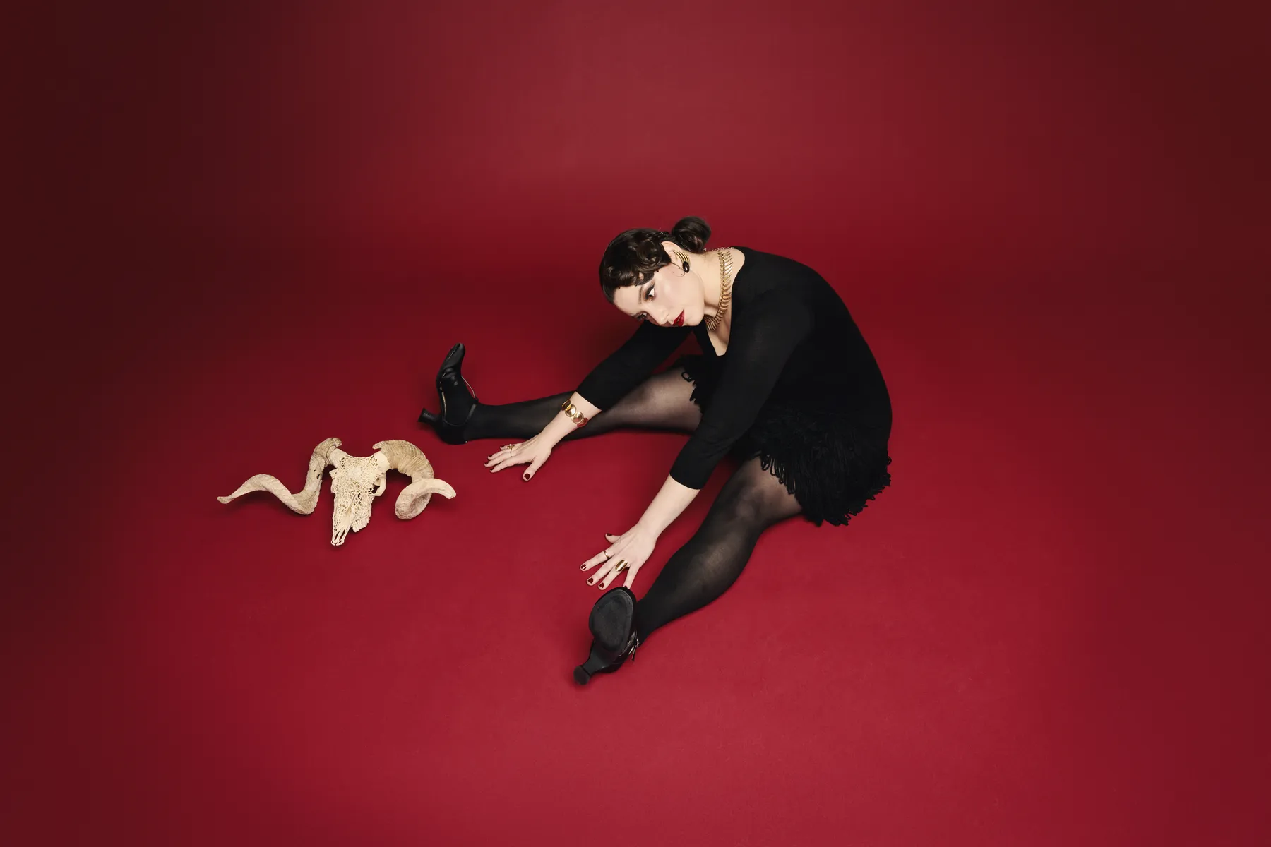 A photo of the artist Gillian Stone showing a woman in a black dress sitting on a red floor with a ram skull beside her