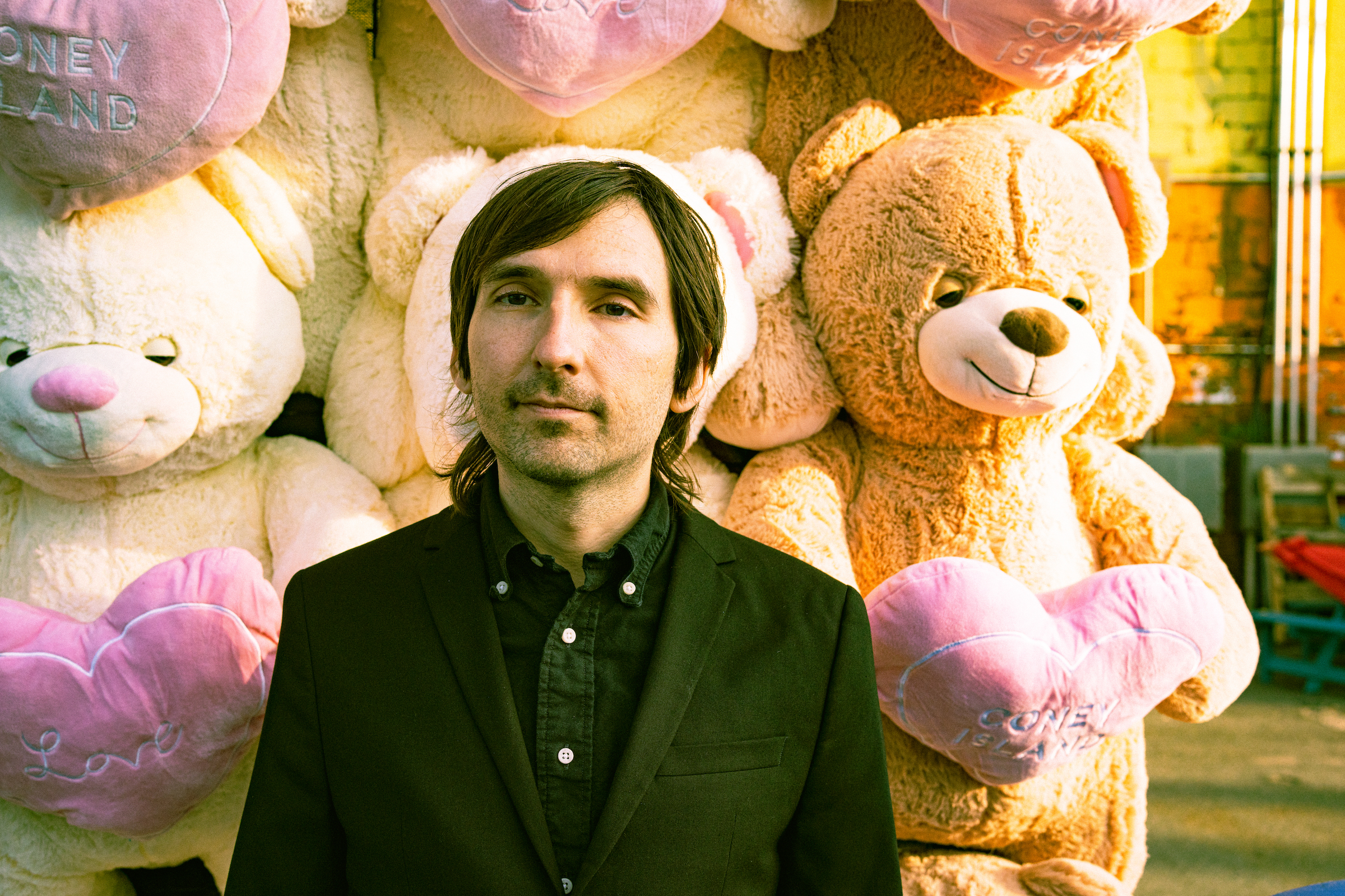 portrait photo of Jordan Lee, aka mutual benefit. He is standing in front of a display of large teddy bears in a fairground
