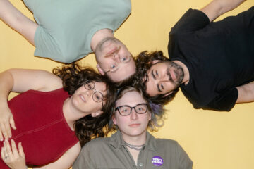 a picture of the band babe report, laying on a yellow floor with their heads touching, looking up at the camera.