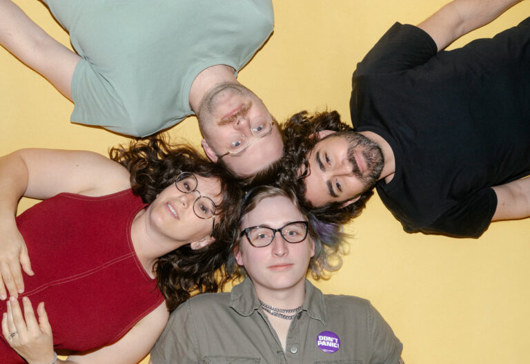 a picture of the band babe report, laying on a yellow floor with their heads touching, looking up at the camera.